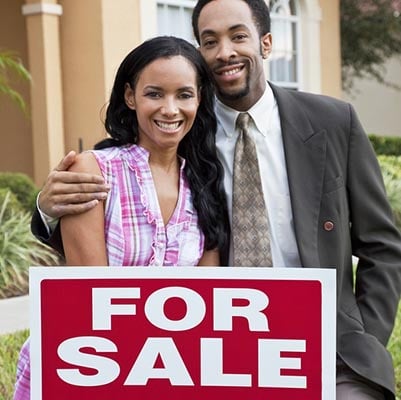 A happy African American man and woman couple outside a large house with a For Sale Sold sign celebrating the purchase of a property